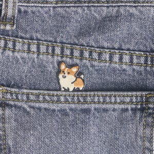 May include: A denim pocket with a small, embroidered corgi patch. The patch features a white and tan corgi design. The pocket is part of a pair of blue jeans, with visible yellow stitching.