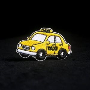 Può includere: Toppa ricamata di un taxi giallo con dettagli neri e bianchi. Il taxi ha il tetto bianco e la scritta "TAXI" è visualizzata sul tetto e sul lato. Sfondo nero.