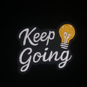 May include: Embroidered patch with the words "Keep Going" in white script on a black background. A yellow lightbulb is to the right of the text. The patch is likely for clothing or accessories.