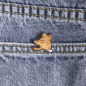 Puede incluir: Primer plano de unos vaqueros azules con un pequeño parche bordado de un gato. El gato es de color naranja y blanco, con contornos negros. El parche está cosido en el bolsillo trasero de los vaqueros.