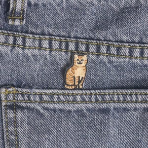 May include: A small, embroidered patch of a tabby cat sitting upright, with a smiling face. The patch is tan and brown, and is placed on the pocket of a pair of blue denim jeans. The jeans have yellow stitching.