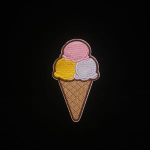 Puede incluir: Parche bordado con un helado de tres bolas. Las bolas son rosa, amarillo y lila, sobre un cono de gofre marrón. El parche tiene un borde marrón y está sobre un fondo negro.