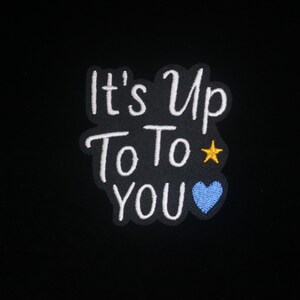 May include: Embroidered patch featuring the text "It's Up To You" in white script on a black background. A yellow star and a blue heart embellish the design. A cheerful accessory for clothing or bags.