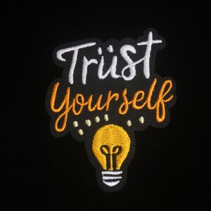 Puede incluir: Parche bordado con las palabras "Trust Yourself" en escritura blanca y naranja. Un gráfico de bombilla amarilla está debajo del texto. El parche está sobre un fondo negro.