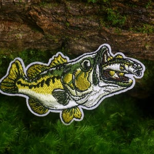 May include: An embroidered patch featuring a large green and yellow bass fish with its mouth open, consuming a smaller fish. The design is rendered in detailed thread work with a white border, showcasing the scales, fins, and eye of the predator.