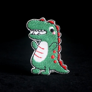 Puede incluir: Parche bordado con un dinosaurio de dibujos animados en tonos verdes, rojos y blancos. El dinosaurio tiene una expresión amigable con una barriga blanca y rayas rojas. El parche es para ropa o accesorios.