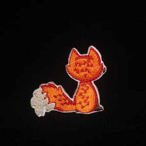 May include: Embroidered patch featuring a cartoon fox in shades of orange, red, and white. The fox is depicted in a seated position with its tail curled up. The patch has a white border and is set against a black background.
