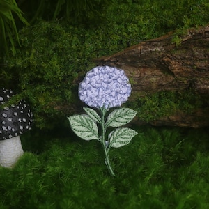 May include: A whimsical scene featuring a light purple hydrangea flower with green leaves, and two decorative mushrooms. One mushroom is black with white spots, and the other is red with white spots. The items are set against a backdrop of green moss and a piece of wood.