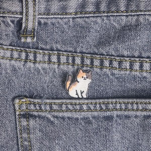 May include: A small, embroidered cat patch in shades of white, orange, and black is sewn onto the pocket of a pair of blue denim jeans. The jeans have yellow stitching and a classic five-pocket design.