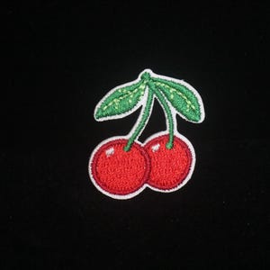 Peut inclure: Écusson brodé représentant deux cerises rouges avec des feuilles et des tiges vertes, sur fond noir. L'écusson a une bordure blanche. Idéal pour personnaliser les vêtements et accessoires.
