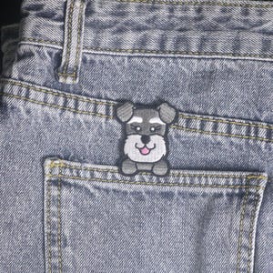 Puede incluir: Vaqueros de mezclilla azul claro con un parche de perro Schnauzer en el bolsillo trasero. El parche es gris, blanco y rosa, con un contorno negro. Los vaqueros tienen costuras amarillas.
