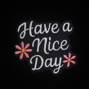 May include: Embroidered patch with the phrase "Have a Nice Day" in white script lettering on a black background. Pink flowers with yellow centers are on either side of the text. A cheerful, positive design.