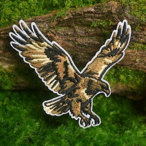 May include: An embroidered patch featuring a golden eagle in flight with wings spread wide. The bird is rendered in shades of brown, tan, and black thread with a white border outline, capturing the eagle in a downward diving motion.