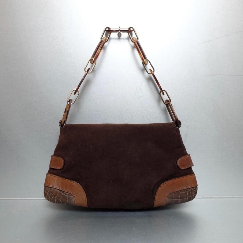 May include: A dark brown suede handbag featuring a structured rectangular body with smooth tan leather accents and embossed crocodile-patterned leather corners. The bag includes a distinctive chain-link handle made of translucent tortoiseshell-style resin.