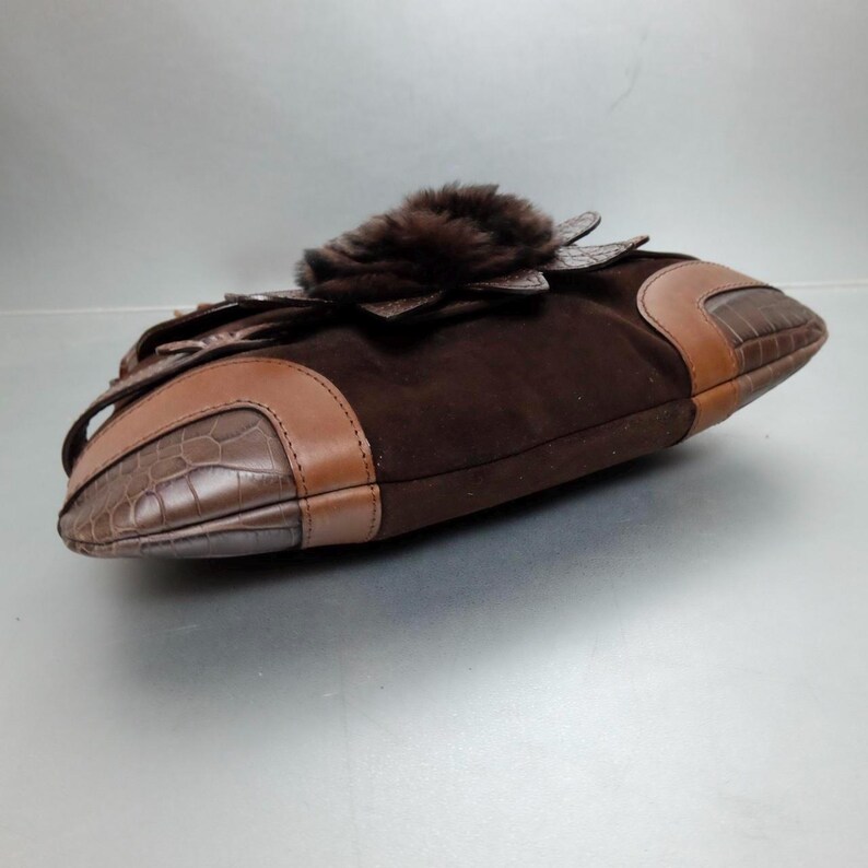 May include: A dark brown suede handbag featuring tan leather trim with a crocodile-embossed texture on the sides. The bag is adorned with a prominent, circular dark brown fur accent centered on the top flap, creating a textured appearance.