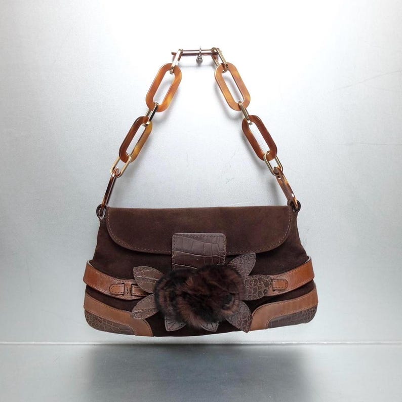 May include: A dark brown suede handbag featuring a flap closure with a central faux fur flower embellishment. The bag includes tan leather trim with a textured pattern and a handle constructed from large, translucent amber-colored plastic chain links.