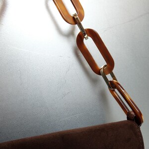 May include: A brown suede handbag featuring a decorative shoulder strap composed of large, oval-shaped, tortoiseshell-patterned resin links connected by small, polished gold-tone metal rings. The strap attaches to the top edge of the bag.