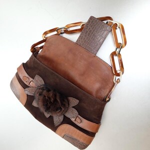 May include: A dark brown suede handbag featuring a smooth leather flap with a crocodile-embossed texture. The bag is adorned with a dark brown faux fur flower applique and includes a decorative chain strap made of large, translucent amber-colored links.