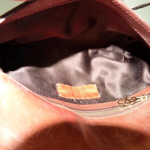 May include: The interior of a brown leather handbag featuring a dark brown satin lining. A rectangular brown leather patch is stitched onto the center of the lining. A gold-toned metal zipper pull with a circular logo is attached to a side pocket.