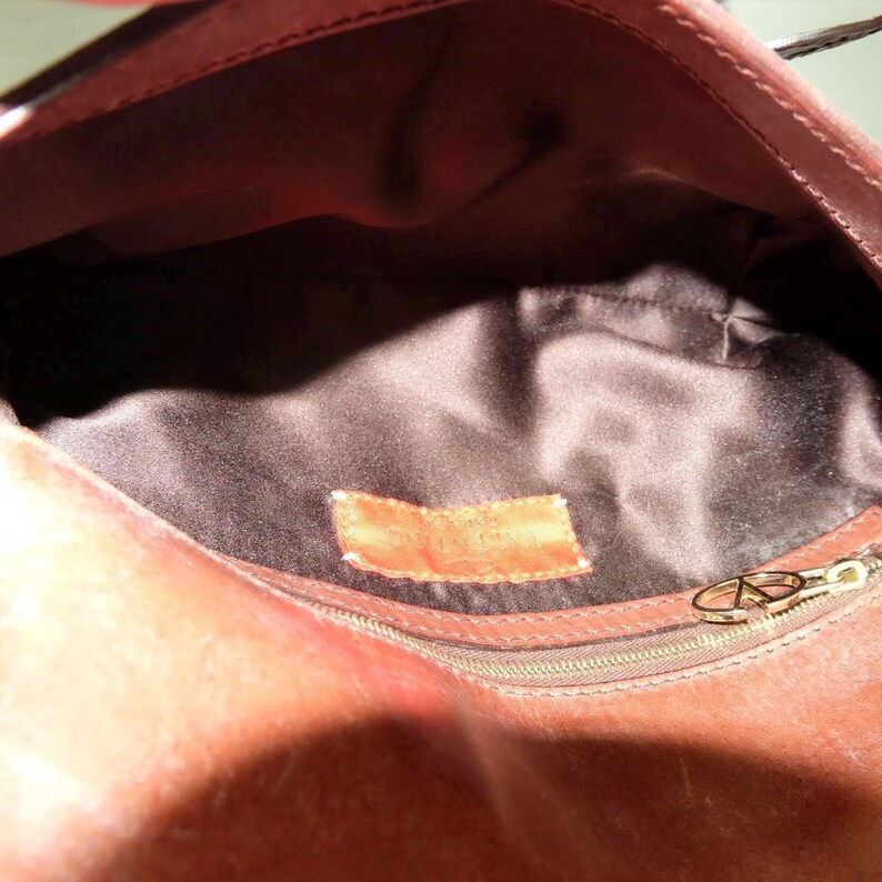 May include: The interior of a brown leather handbag featuring a dark brown satin lining. A rectangular brown leather patch is stitched onto the center of the lining. A gold-toned metal zipper pull with a circular logo is attached to a side pocket.