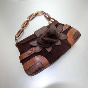 May include: A dark brown suede handbag featuring a textured brown leather trim with a crocodile pattern. The bag includes a decorative dark brown fur flower applique on the front flap and a handle made of translucent amber-colored plastic chain links.