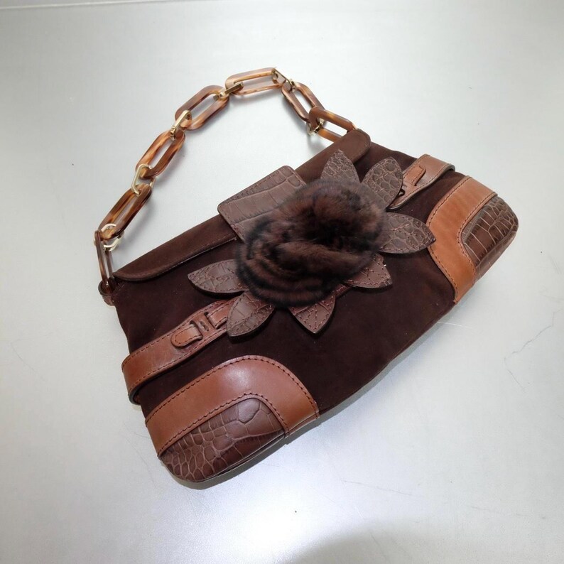May include: A dark brown suede handbag featuring a textured brown leather trim with a crocodile pattern. The bag includes a decorative dark brown fur flower applique on the front flap and a handle made of translucent amber-colored plastic chain links.