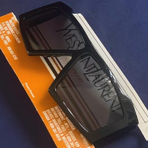 May include: Black square-framed sunglasses with the brand name "YVES SAINT LAURENT" printed on the lenses. The sunglasses rest on an orange and white product information card. The text "Frame France" is visible.