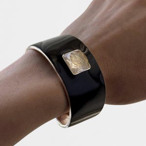 May include: A wide, smooth black cuff bracelet featuring a single, square-cut, translucent yellow gemstone set in a silver-toned metal frame. The bracelet has a polished, reflective surface and a metallic interior lining, creating a bold contrast.