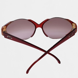 May include: A pair of sunglasses with deep red frames and gradient lenses. The temples feature a silver-toned metallic logo emblem on each side. The frames have a distinct curved shape, and the temples are crossed behind the lenses.