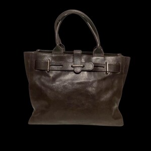 May include: A dark brown leather handbag featuring two rounded top handles and a horizontal strap across the front. The strap is secured with a central tab and includes two vertical metal accents on either side, providing a structured appearance.