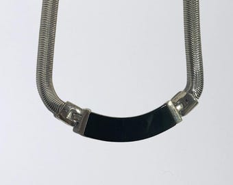 May include: A silver-toned snake chain necklace featuring a smooth, curved black rectangular centrepiece. The metal chain has a flexible, tightly woven texture that connects to the central black element with polished silver-toned metal hinges.