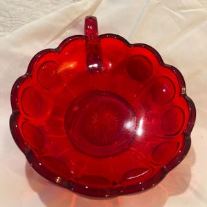 Könnte beinhalten: Eine leuchtend rote Glasschale mit gewelltem Rand und kleinem Griff. Die Schale hat ein zentrales Design und kreisförmige Muster am Rand. Dieses dekorative Stück eignet sich zum Servieren oder Ausstellen.