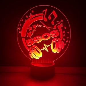 May include: A red LED night light with the text "5SOS" in a circular design. The design includes musical notes, stars, and a stylized sound wave. The base is black.