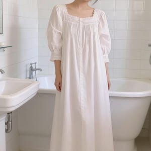 May include: A white, long-sleeved nightgown with a square neckline and lace trim. The nightgown features pleats on the bodice and cuffs, and a button-up front. The garment is made of a soft, flowing fabric.