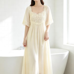 May include: A floor-length cream-coloured nightgown featuring a square neckline with intricate floral lace detailing, short flutter sleeves with ruffled edges, and a flowing, lightweight skirt that falls from a gathered bodice.