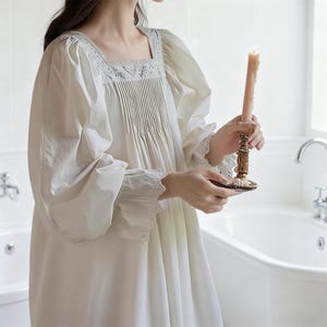 May include: A cream-colored nightgown with a square neckline and pleated bodice. The long sleeves have ruffled cuffs with eyelet detailing. A lit candle in a gold-colored candlestick is held in the hands.