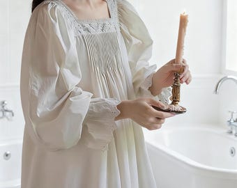 Può includere: Una camicia da notte color crema con scollo quadrato e corpetto plissettato. Le maniche lunghe hanno polsini arricciati con dettagli traforati. Si tiene una candela accesa in un candelabro dorato.