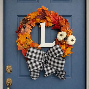 May include: A seasonal wreath featuring autumnal leaves in shades of orange and red, with white pumpkins and a black and white gingham bow. A white letter "L" is displayed. The wreath is on a blue door.