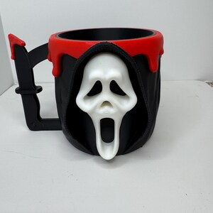 May include: A black and red Halloween-themed mug featuring a white Ghostface mask design. The mug has a black handle shaped like a knife and a red rim that appears to be dripping. The mug is a unique and spooky addition to any Halloween collection.