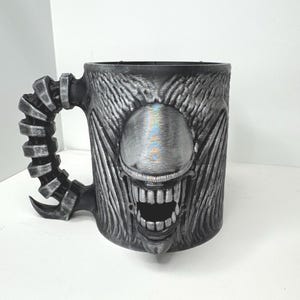 May include: A silver and black ceramic mug with a monster face design. The mug features a detailed, textured surface, a large open mouth, and a handle shaped like a spine. The mug is approximately 4 inches tall.