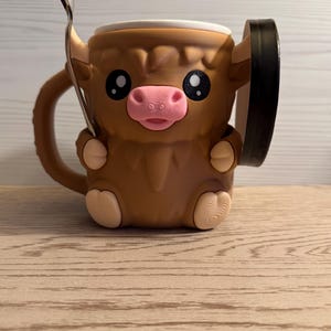 May include: A brown ceramic mug shaped like a Highland cow, featuring a pink nose, large black eyes, and a white rim. A silver spoon rests inside the mug. The mug has a handle and a black accent on the side.