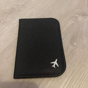 May include: Black passport holder with a small white airplane icon in the corner. The holder is rectangular with rounded edges and a textured surface. The background is a light wood grain.