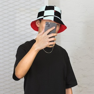 May include: Reversible bucket hat. One side features a black and white chequered pattern, the other is solid red. A small red heart detail is on the chequered side. The hat is worn by a person wearing a black t-shirt.