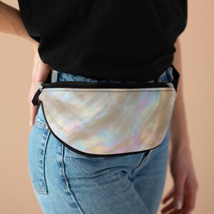 May include: A pearlescent bum bag with a black zip and trim. The bag has a curved shape and is worn around the waist. The iridescent material reflects a range of colours, including pink, yellow, and blue.