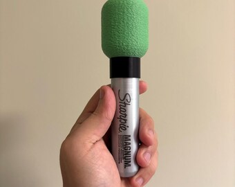 May include: A silver Sharpie Magnum permanent marker with a green foam cover on top. The marker is held in a person's hand. The text "Sharpie MAGNUM" is printed on the marker.