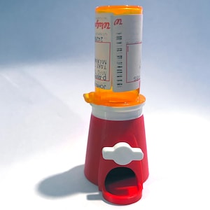Può includere: Un distributore di pillole rosso e bianco con un flacone di pillole trasparente sopra. Il flacone di pillole ha un tappo arancione e un'etichetta con testo. Il distributore ha una fessura per erogare le pillole.