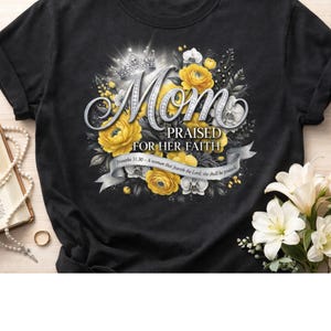 Puede incluir: Camiseta negra con un diseño floral. El diseño incluye la palabra "Mom" en plata, una corona y rosas amarillas. El texto "PRAISED FOR HER FAITH" está debajo, con una cinta que dice "Proverbios 31:30 - La mujer que teme al Señor, será alabada."