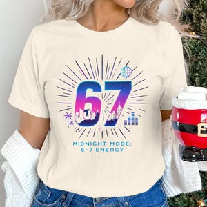 May include: A cream-coloured t-shirt displaying a colourful graphic of the number '67' in blue and purple, with radiating lines and cityscapes. The text below reads 'Midnight Mode: 6-7 Energy'. A festive mug is held in the hand.