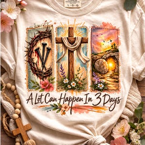 May include: Cream-colored t-shirt with a graphic design featuring a cross, crown of thorns, and empty tomb. The text "A Lot Can Happen In 3 Days" is printed below the images. The shirt is adorned with floral accents.
