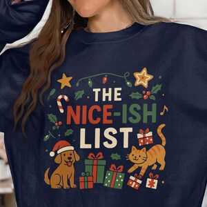 May include: Navy blue sweatshirt featuring a Christmas-themed design. The text "THE NICE-ISH LIST" is displayed with illustrations of a dog in a Santa hat, a cat, and wrapped presents. Festive stars, holly, and candy canes complete the design.